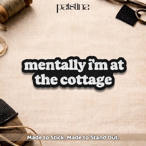 May include: A black and white embroidered patch with the text "mentally I'm at the cottage." The patch is on a beige fabric surface. The brand name "patstina" is at the top. The text "Made to Stick. Made to Stand Out." is at the bottom.