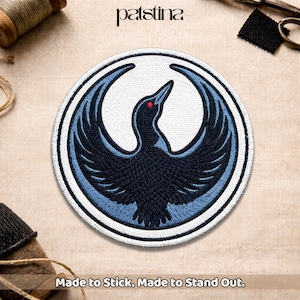 May include: Circular embroidered patch featuring a stylized black bird with outstretched wings, a red eye, and a blue outline, set against a white background. The patch is bordered by a blue and white ring. Text at the bottom reads "Made to Stick. Made to Stand Out."