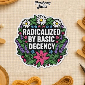May include: Embroidered patch featuring the text "RADICALIZED BY BASIC DECENCY" in white, encircled by a wreath of green leaves, white daisies, and pink and purple flowers. Set against a beige fabric backdrop.