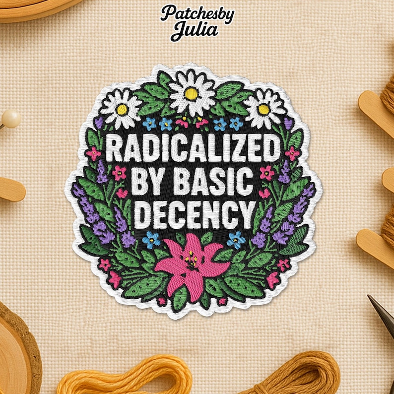 May include: Embroidered patch featuring the text "RADICALIZED BY BASIC DECENCY" in white, encircled by a wreath of green leaves, white daisies, and pink and purple flowers. Set against a beige fabric backdrop.