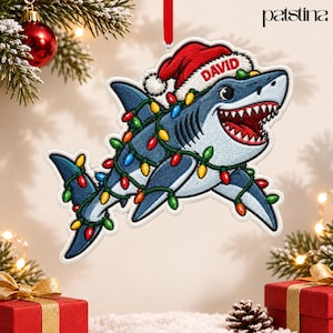 May include: A shark-shaped Christmas ornament featuring a blue shark wearing a red Santa hat with the name DAVID printed on the brim. The shark is wrapped in a string of colorful Christmas lights with red, green, yellow, and blue bulbs.