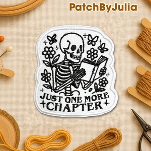 May include: A white embroidered patch featuring a skeleton reading a book, surrounded by flowers and butterflies. The text "JUST ONE MORE CHAPTER" is below the skeleton. The patch is on a beige background with sewing supplies.