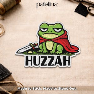 May include: Embroidered patch of a green frog in a red cape, holding a sword, with the word "Huzzah" in white. The patch is on a beige background. The brand name "patsina" is at the top.