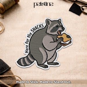 May include: An embroidered patch featuring a gray and black raccoon holding a tan sandwich. The text "Here for the SNACKS" is arched above the raccoon in black lettering. The patch has a white border and is designed for fabric application.