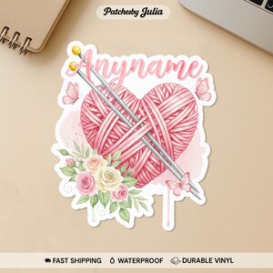 May include: A sticker featuring a pink heart made of yarn, with knitting needles, butterflies, and flowers. The word "Anyname" is written in pink script across the top. The sticker has a watercolor effect.
