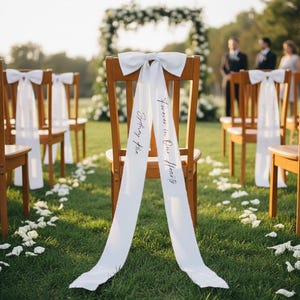 Op de afbeelding: Houten stoelen met witte strikken en stromende linten zijn opgesteld voor een buitenceremonie. De linten op de stoelen hebben de tekst "Forever in Our Hearts" en "Sydney Rae". Witte bloemblaadjes zijn verspreid over het groene gras.