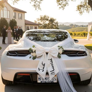 Op de afbeelding: Een witte auto versierd voor een bruiloft met een grote strik en sleepdoek. De strik is versierd met bloemen en de woorden "Just Married" staan erop gedrukt. Een kentekenplaat luidt "MR & MRS _024".