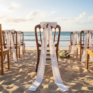 May include: Wooden chairs with white bows and flowing ribbons are arranged on a sandy beach. The ribbons have text that reads "Forever in Our Hearts" and "Sydney Rae". A bouquet of white flowers sits near the chairs, with the ocean in the background.