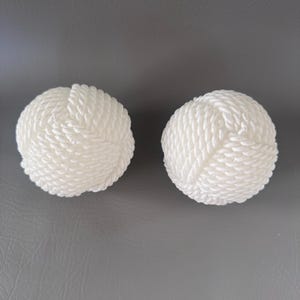 May include: Two white decorative spheres with a rope-like texture. The spheres are round and appear to be made of tightly wound, off-white rope. They are placed on a gray surface.