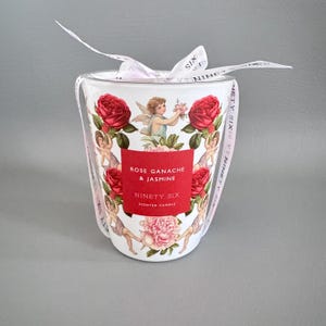 Puede incluir: Una vela perfumada blanca con una etiqueta roja que dice "Rose Ganache & Jasmine" y "Ninety Six". La vela est&aacute; decorada con rosas rojas, querubines y un lazo rosa.