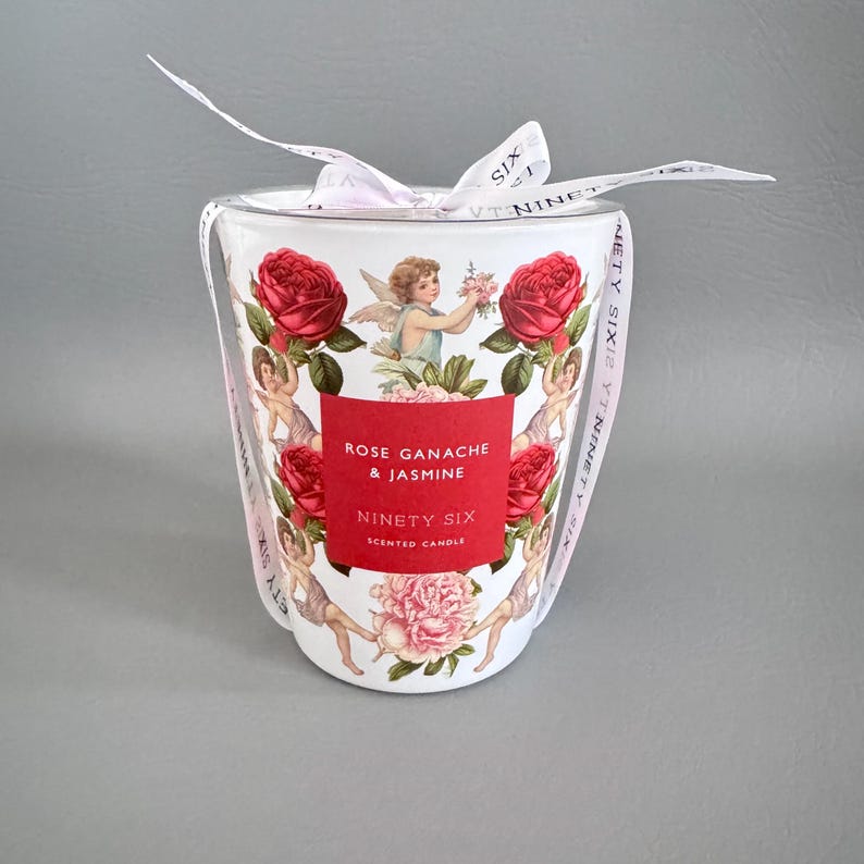Puede incluir: Una vela perfumada blanca con una etiqueta roja que dice "Rose Ganache & Jasmine" y "Ninety Six". La vela est&aacute; decorada con rosas rojas, querubines y un lazo rosa.