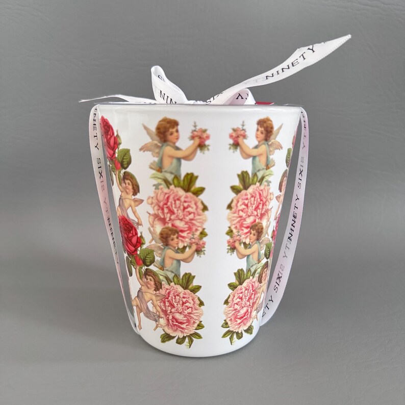 Puede incluir: Taza de cer&aacute;mica blanca con un dise&ntilde;o floral y de querubines. La taza est&aacute; adornada con peon&iacute;as rosas, rosas rojas y querubines sosteniendo flores. Una cinta blanca con el texto "NINETY SIX" est&aacute; atada alrededor de la taza.