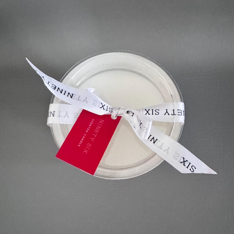 Puede incluir: Una vela perfumada blanca y redonda en un recipiente de pl&aacute;stico transparente. Una cinta blanca con el texto "SIXTY TWO YT NINET" est&aacute; atada alrededor del recipiente. Una etiqueta roja con el texto "NINETY SIX SCENTED CANDLE" est&aacute; sujeta a la cinta.