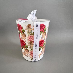 Puede incluir: Un vaso cil&iacute;ndrico blanco adornado con un patr&oacute;n repetido de querubines, rosas rojas y peon&iacute;as rosas. Una cinta blanca con el texto "NINETY SIX" est&aacute; atada alrededor de la parte superior. El vaso es probablemente para bebidas.