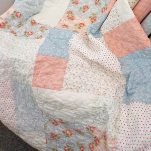 Może przedstawiać: Kołdra patchworkowa z różnymi wzorami kwiatowymi w odcieniach różowego, niebieskiego i białego. Kołdra jest wykonana z bawełny i ma pikowaną fakturę.