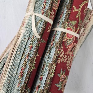 May include: Two stacks of fabric strips tied with white ribbon. The fabric features a deep red background with intricate floral patterns in shades of light blue, green, and tan. Each strip has pinked, zigzag edges, creating a textured appearance.