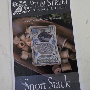 May include: A cross stitch pattern sampler featuring a pig with a blue and green floral design. The pattern is titled "Snort Stack" and is from Plum Street Samplers, original designs by Paulette Stewart.