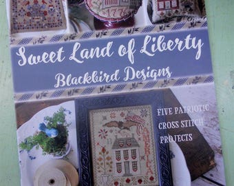 May include: Book cover featuring a blue and white striped background with the title "Sweet Land of Liberty" and "Blackbird Designs". The cover also includes five cross stitch projects with patriotic themes, including a house with an American flag, a bird, and a banner that says "America".