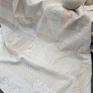 May include: A white patchwork quilt with a variety of textures and patterns, including small floral designs, dots, and stripes. The quilt is folded and draped over a surface, with a white knitted pumpkin resting on top.