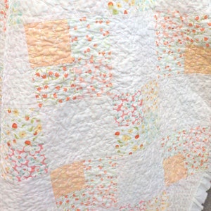 Könnte beinhalten: Eine weiße Patchwork-Steppdecke mit gerüschtem Rand. Die Steppdecke zeigt Quadrate mit Blumenmustern in Pfirsich, Rosa, Gelb und Grün auf weißem Hintergrund. Die Steppdecke hat eine gesteppte Textur.