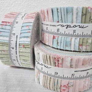 May include: Rolled fabric bundles in pastel colors, including pink, blue, and green, are secured with white measuring tape-style ribbons. The ribbons feature the word "moda" and the website address www.modafabrics.com.