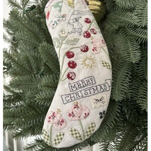 May include: Embroidered Christmas stocking with floral and berry designs. The stocking features the text "MERRY CHRISTMAS" and the number "25". The stocking is decorated with a variety of embroidered elements, including flowers, berries, and a dog.
