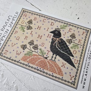 May include: A cross-stitch pattern titled "Miss Raven's Pumpkin Sampler." The design features a black raven perched on a pumpkin, surrounded by acorns and the alphabet. The pattern is on a cream-colored background with a black border.