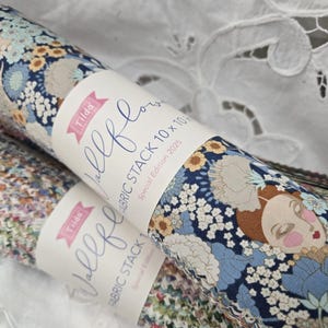 May include: Two rolls of patterned fabric, featuring floral designs and a label reading "Tilda Vallel Flowers Fabric Stack 10 x 10". The fabric displays a woman's face and flowers in blue, white, and brown hues.