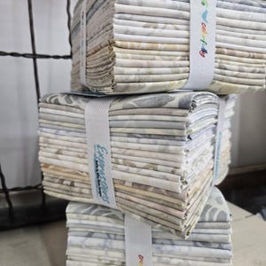 May include: Three stacks of fabric bundles, each secured with a white ribbon. The fabric features a soft, floral pattern in shades of grey, blue, and cream. The ribbons have text that reads "Expressions" and "create colorfully".