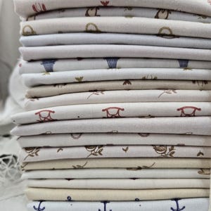 May include: A pile of folded fabric squares in cream and beige tones. Each piece is decorated with a different pattern, such as anchors, baskets, and floral motifs. The fabric looks soft and light, ideal for crafting and sewing.