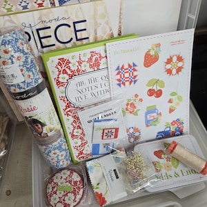 May include: A clear plastic storage container filled with quilting supplies. Items include fabric rolls, books, stickers, quilt labels, and a small gold chain. The items have floral and geometric patterns in red, blue, and white.