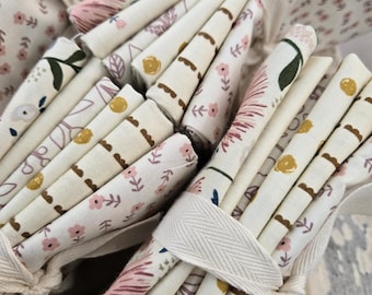 May include: A stack of fabric squares with various floral and geometric patterns in shades of pink, brown, and yellow on a white background. The fabric is tied together with a white cotton ribbon.