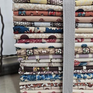 May include: A pile of patterned fabric squares in shades of blue, yellow, red, and brown, secured with a grey ribbon. The fabrics display floral and geometric patterns, giving a vintage feel. The stack is neatly arranged, highlighting the diverse patterns.