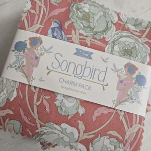 May include: A stack of fabric squares with a floral pattern in shades of coral, green, and cream. A label wrapped around the squares reads "Songbird Charm Pack" with illustrations of a blue bird and a woman. The fabric is likely for quilting or crafting.