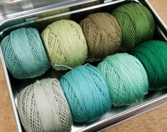 May include: A metal tin with a clear lid containing eight balls of green yarn in various shades of green.