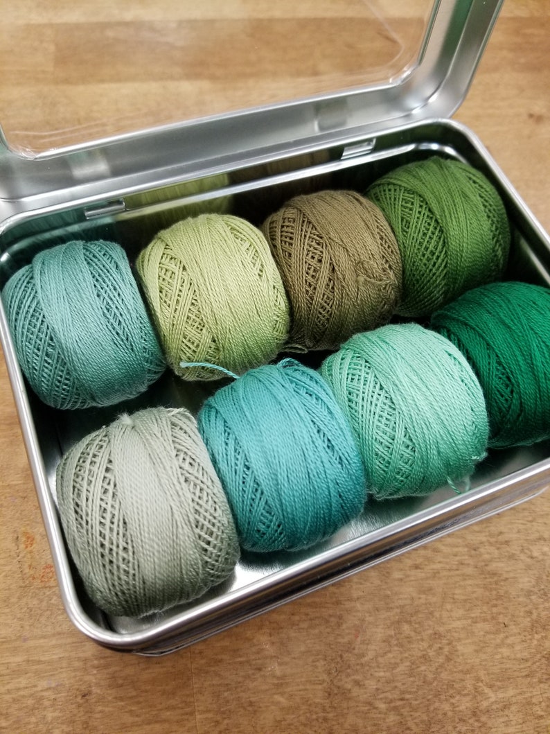 May include: A metal tin with a clear lid containing eight balls of green yarn in various shades of green.