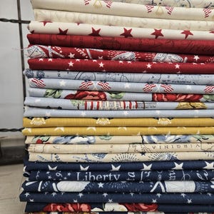 Könnte beinhalten: Ein Stapel Stoffstücke mit patriotischen Designs in Rot, Weiß und Blau. Die Muster umfassen Sterne, Karten und den Schriftzug "Liberty". Die Stoffe sind gestapelt und zeigen eine Vielzahl von Drucken für Bastelprojekte.