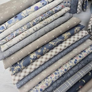 May include: A collection of fabric pieces in shades of blue, grey, and beige. The fabrics display floral, checked, and patterned designs. Some fabrics have a textured appearance, while others are smooth. The fabrics are neatly arranged, showcasing a variety of patterns.