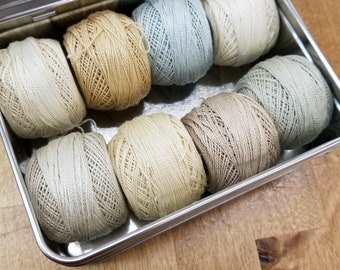 May include: A metal tin filled with eight balls of yarn in various shades of beige, cream, and light blue.
