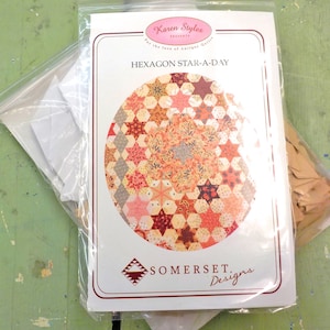 May include: A white paper package with a quilting pattern titled "Hexagon Star-A-Day" by Karen Styles. The package includes a colorful quilt design, a white paper template, and a brown paper template.