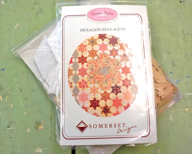 May include: A white paper package with a quilting pattern titled "Hexagon Star-A-Day" by Karen Styles. The package includes a colorful quilt design, a white paper template, and a brown paper template.