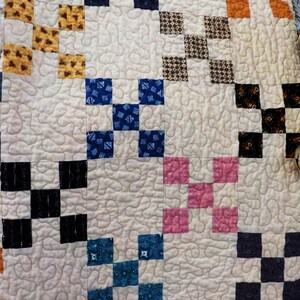 May include: Close-up of a handmade quilt with a white background and a repeating pattern of colorful squares. The squares are in shades of blue, orange, pink, black, brown, and yellow. The quilt is machine quilted with a meandering stitch.