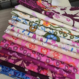 Peut inclure: Une pile de tissus pliés avec divers motifs floraux et fantaisistes. Les tissus présentent une gamme de couleurs, notamment du rose, du violet, du bleu et du crème. Certains motifs comprennent des fleurs, des lapins et des motifs abstraits. Une boucle blanche est visible en haut.