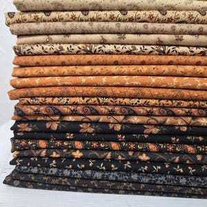 May include: A vertical stack of folded fabric pieces featuring a gradient of colours from cream and beige to warm orange, brown, and deep black. Each layer displays a unique repeating pattern of small floral, botanical, and paisley motifs.