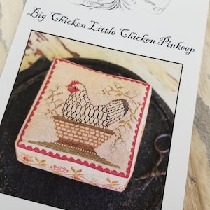 May include: Counted cross-stitch pattern for a project titled "Big Chicken Little Chicken Pinkeep". The pattern features a white chicken sitting in a brown basket with green leaves and white flowers. The pattern is surrounded by a red and gold border.