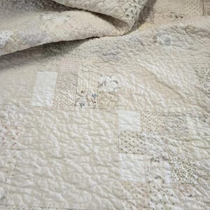 May include: A white quilt with a patchwork design made from various shades of white and cream fabrics. The quilt is made of squares and rectangles, and the fabric has a variety of textures, including smooth, textured, and patterned.