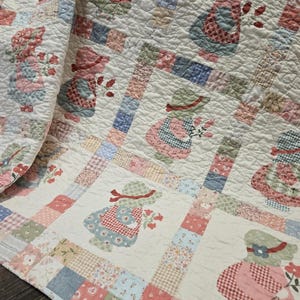 May include: A patchwork quilt featuring repeating appliqued figures of children holding flowers. The quilt has a cream background with squares of various pastel fabrics. The figures are in dresses and bonnets, with red, pink, blue, and green accents.