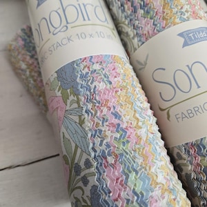 May include: Rolled fabric stacks with a floral pattern in pastel colors. The fabric is labeled "Songbird Fabric Stack 10 x 10 in" and features a design with birds and flowers. The fabric has a zig-zag edge.