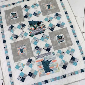 May include: A white quilt with a blue and grey border. The quilt features a patchwork design with a central panel depicting a map of Minnesota with the word "Minnesota" written on it. The map is surrounded by smaller squares with various designs.