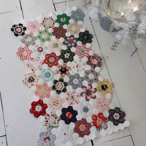May include: A white fabric runner with a Christmas theme. The runner is made of hexagonal fabric pieces in various colors and patterns, including red, green, blue, and white. The runner is decorated with snowflakes and other Christmas motifs.